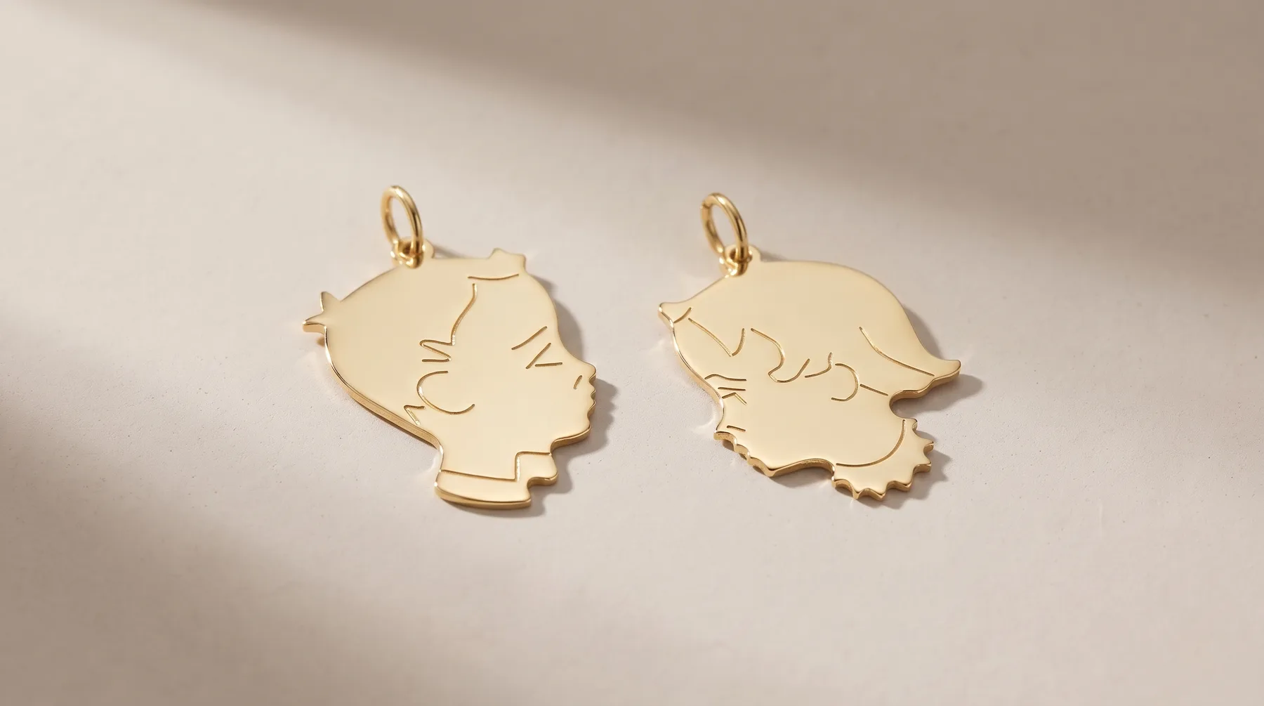 Two gold pendant charms with small loops, each featuring a minimalist line-drawn profile of a child's head facing opposite directions, displayed on a neutral background.