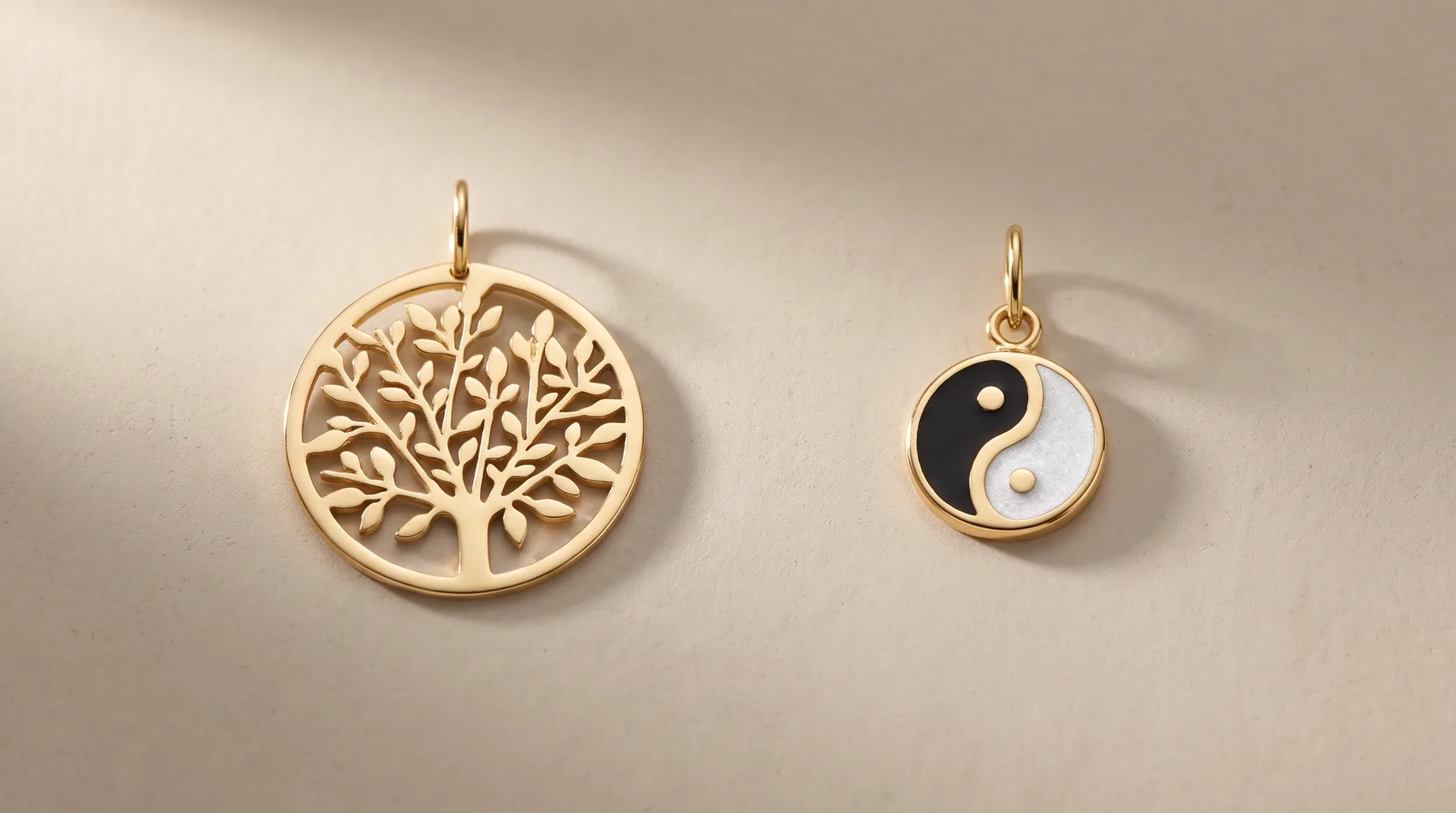 Two gold pendant charms on a beige surface: a larger circular pendant with a tree design and a smaller circular pendant with a yin-yang symbol in black and white enamel.