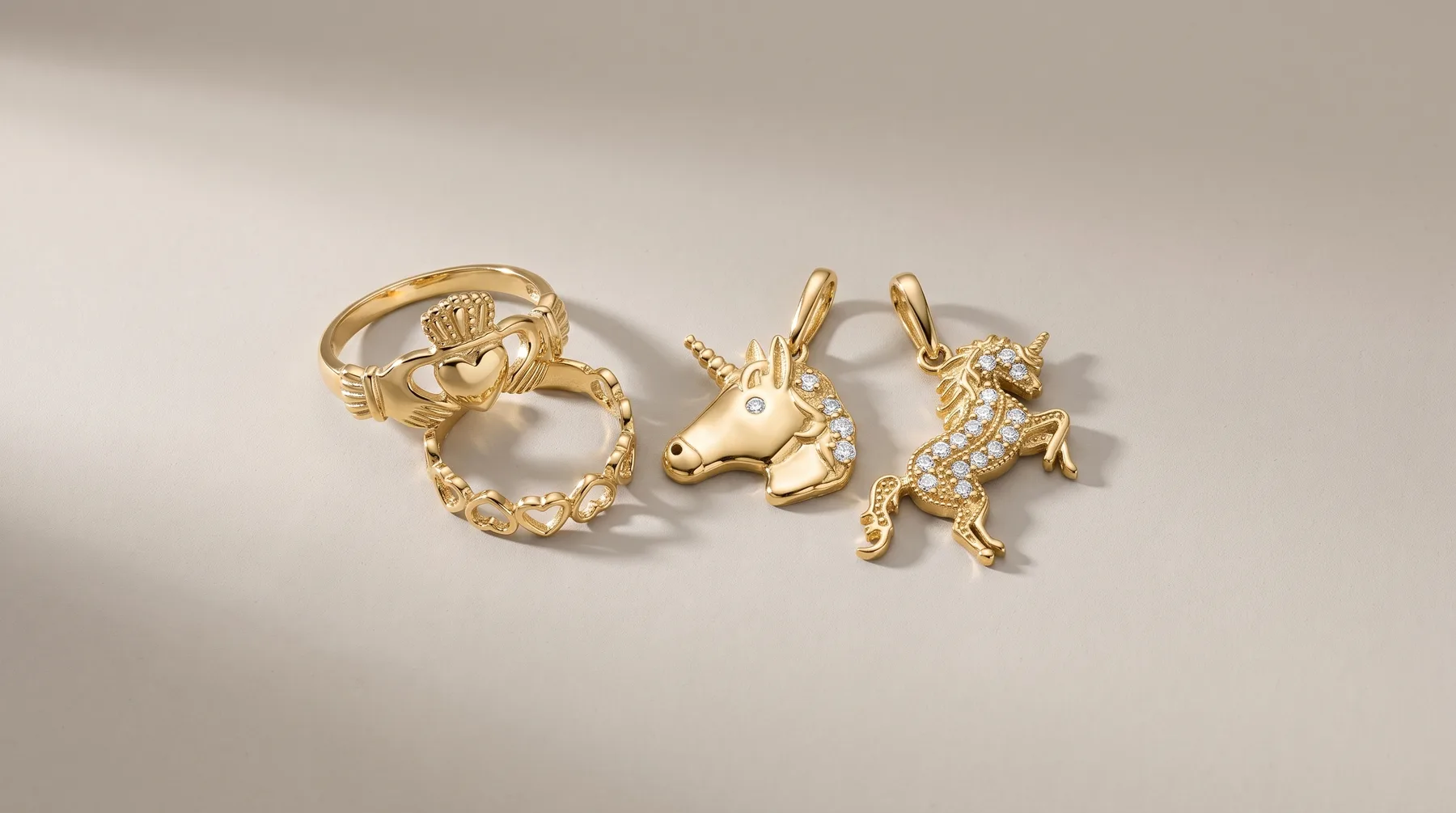 Three gold rings and pendants arranged on a light surface: a claddagh-style ring, a textured band ring, a rabbit charm, and a goat charm with cubic zirconia stones.