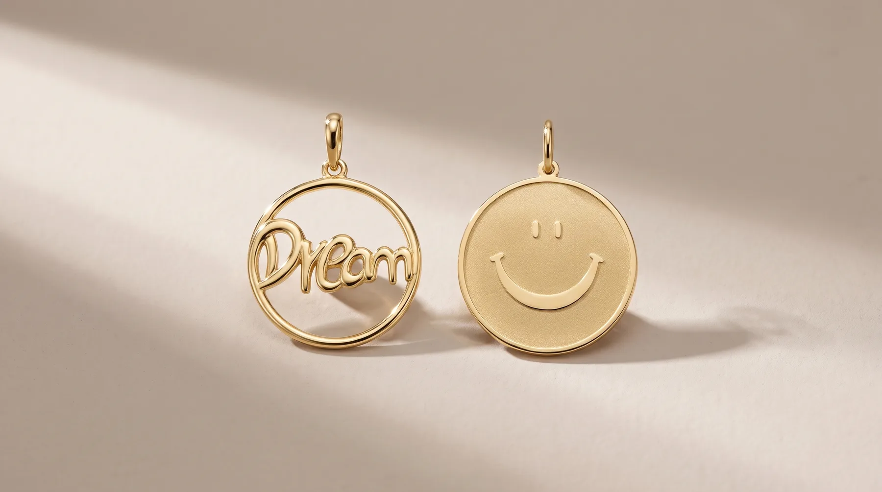 Two gold circular pendants displayed against a neutral background: one featuring the word "Dream" in an open circle design, the other a solid disc with an engraved smiley face.