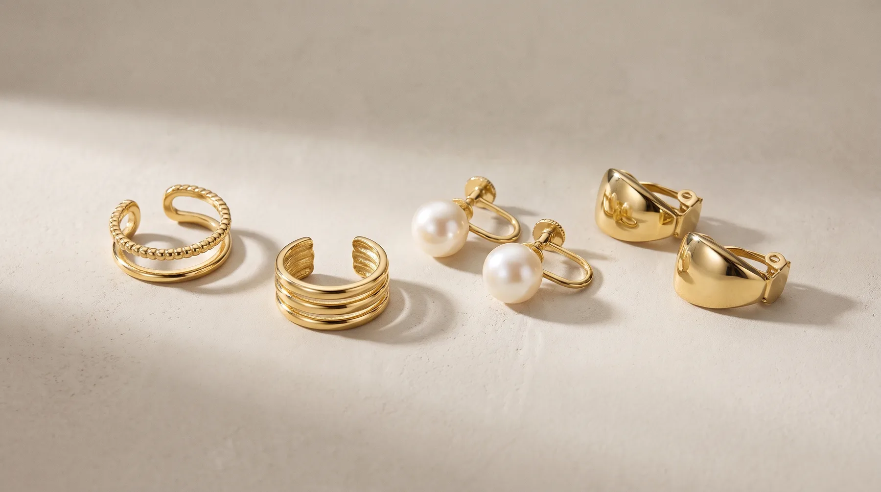 Five gold-toned rings and earrings arranged on a white surface, including textured bands, pearl stud earrings, and chunky polished styles.
