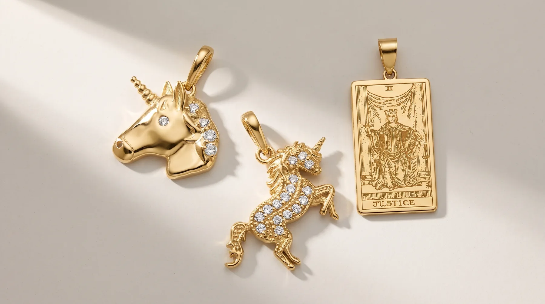 Three gold pendants with bail loops arranged on a white surface: a unicorn head with diamond accents, a prancing horse with pavé diamonds, and a tarot card design labeled Justice.