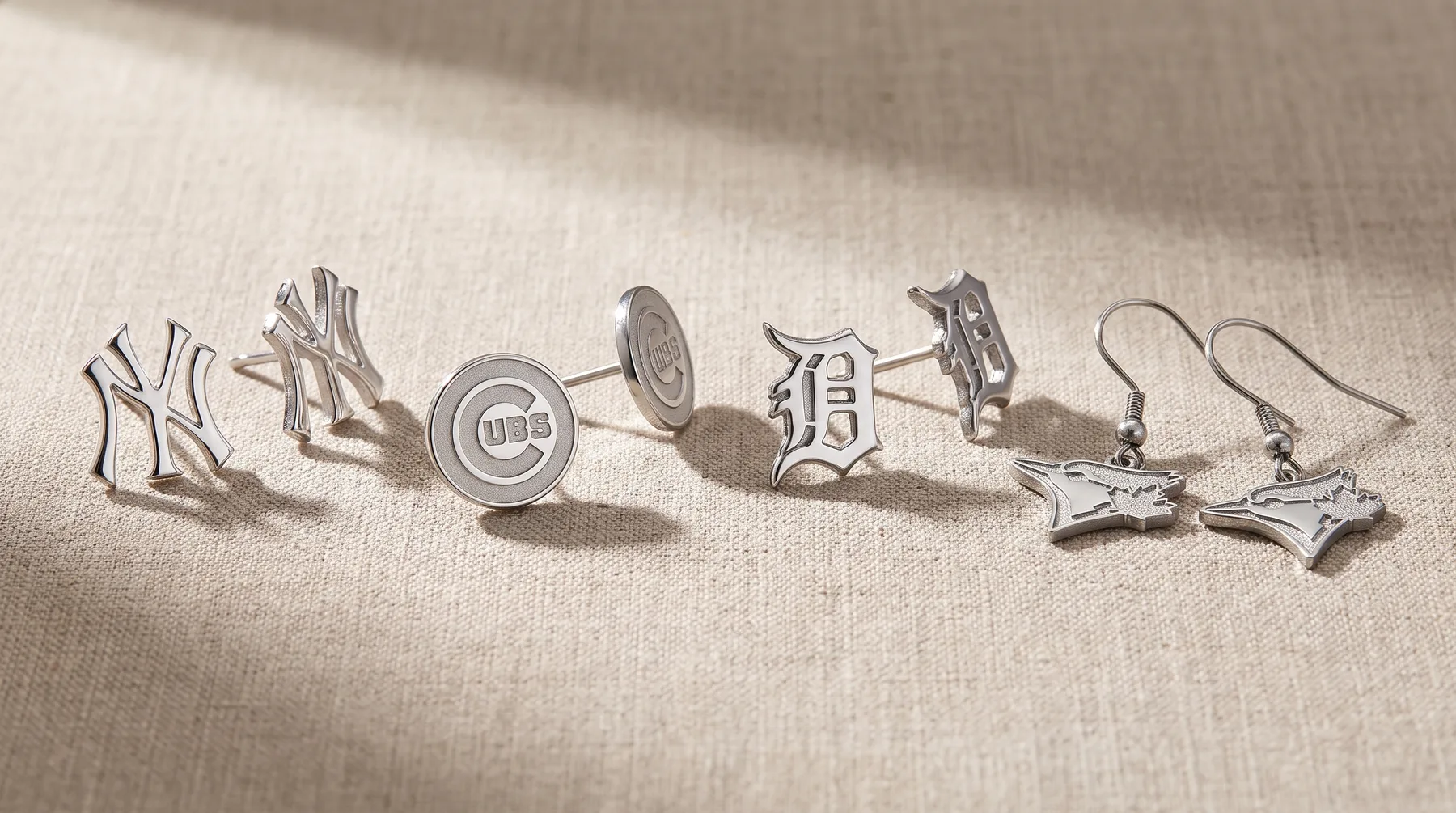Five pairs of silver-toned metal cufflinks and earrings displayed on a light linen fabric, including baseball team logos and bow-tie designs.