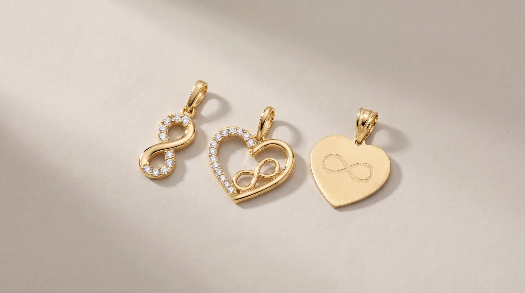 Three gold pendants with infinity symbol designs arranged on a light surface, including one with diamonds, one heart-shaped with diamonds, and one plain heart-shaped pendant.
