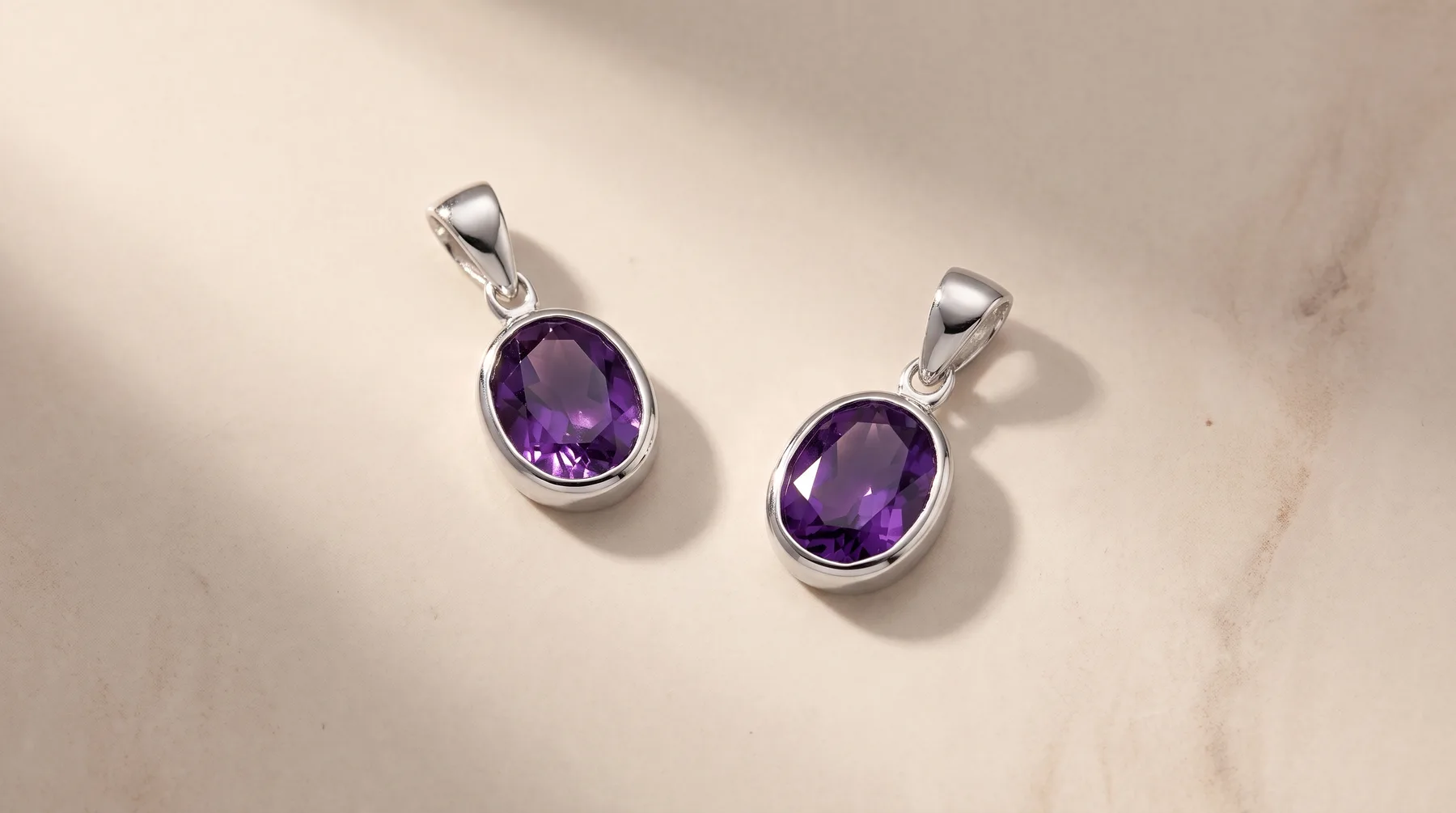 Two silver pendants with oval purple amethyst stones set in bezels, displayed on a light marble surface with natural shadows.