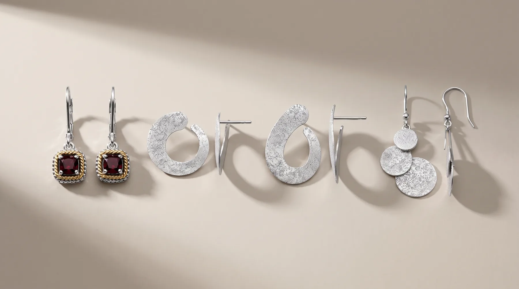 Six pairs of silver and gemstone earrings displayed in a row on a neutral surface, including drop styles with red stones, textured circular designs, and multi-disc pendants.