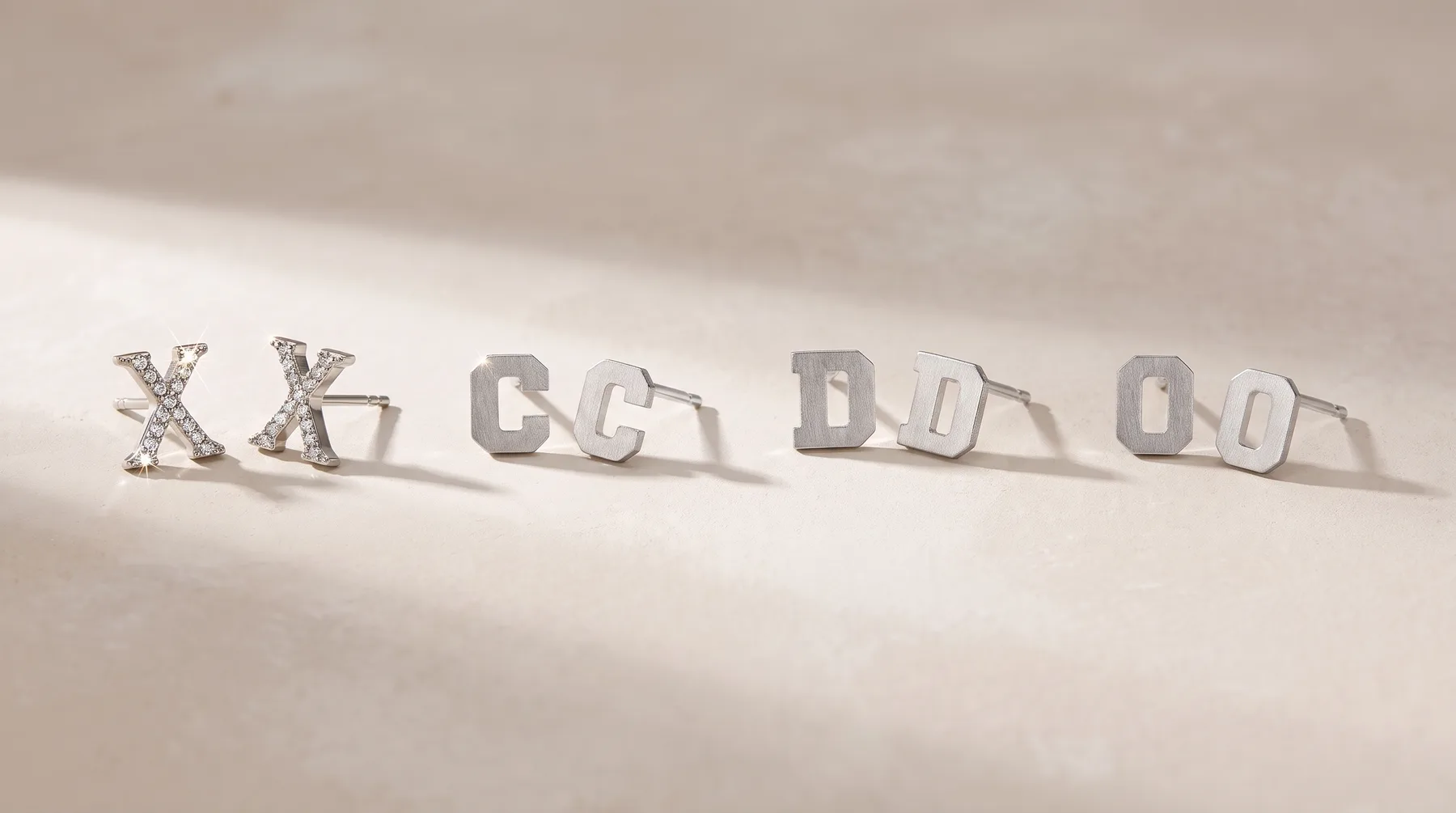 Silver stud earrings with X design and matching silver stud earrings with geometric shapes arranged in a line on a beige surface.