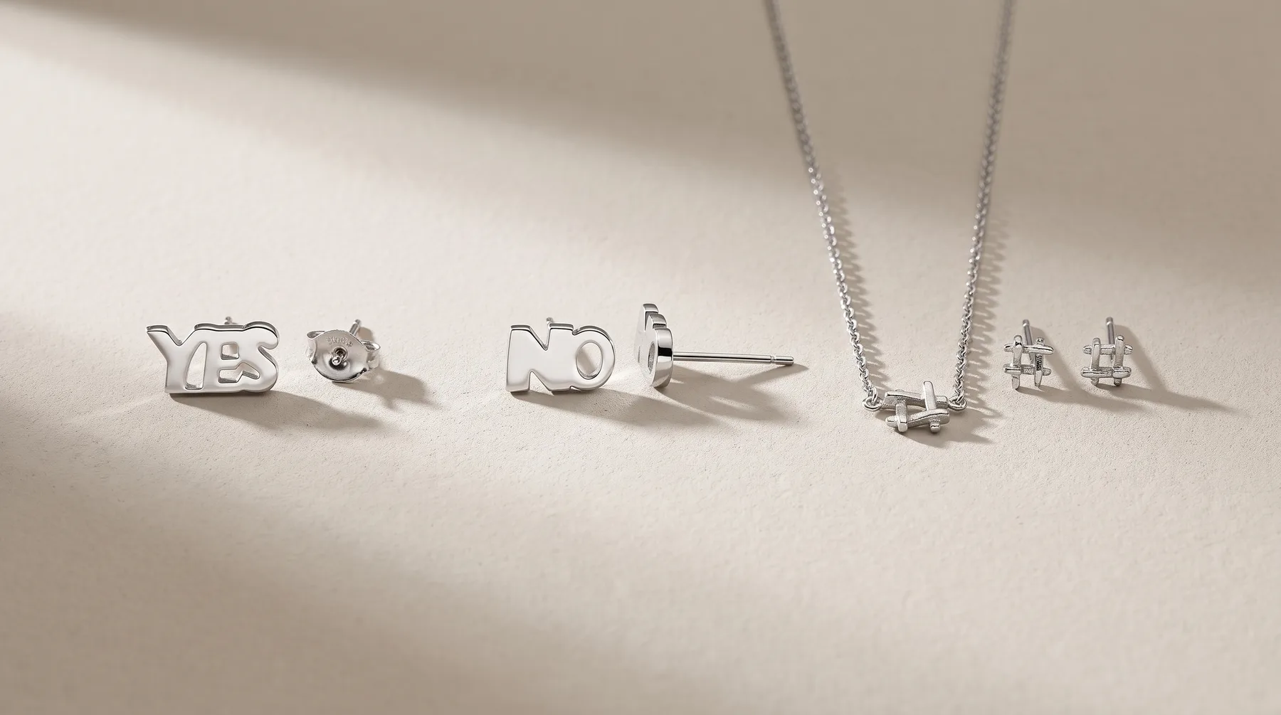 Silver jewelry pieces arranged on a white surface: "YES" and "NO" stud earrings on the left, a "NO" stud earring in the center, a necklace with a small pendant and chain on the right, and cubic zirconia stud earrings.