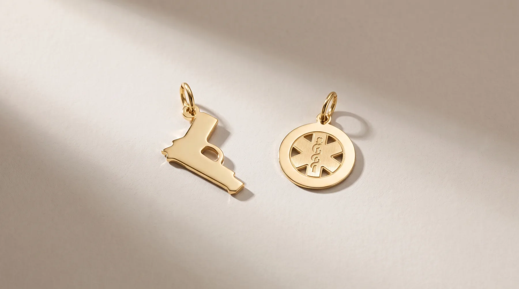Two gold charms with jump rings cast in relief: a shoe-shaped charm on the left and a circular medical symbol charm on the right, displayed on a white surface with dramatic shadow.