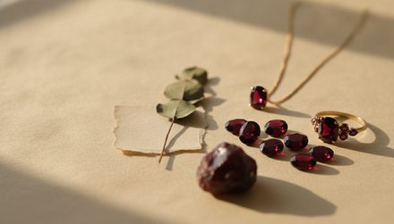 Deep red garnet gemstones, both raw and faceted, arranged on beige paper beside a gold ring, pendant necklace, and small eucalyptus leaves.