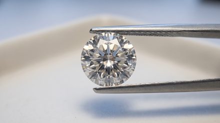 A round brilliant diamond held face-up in steel tweezers, its facet pattern filling the frame, with a small dark crystal inclusion visible just off centre beneath the table