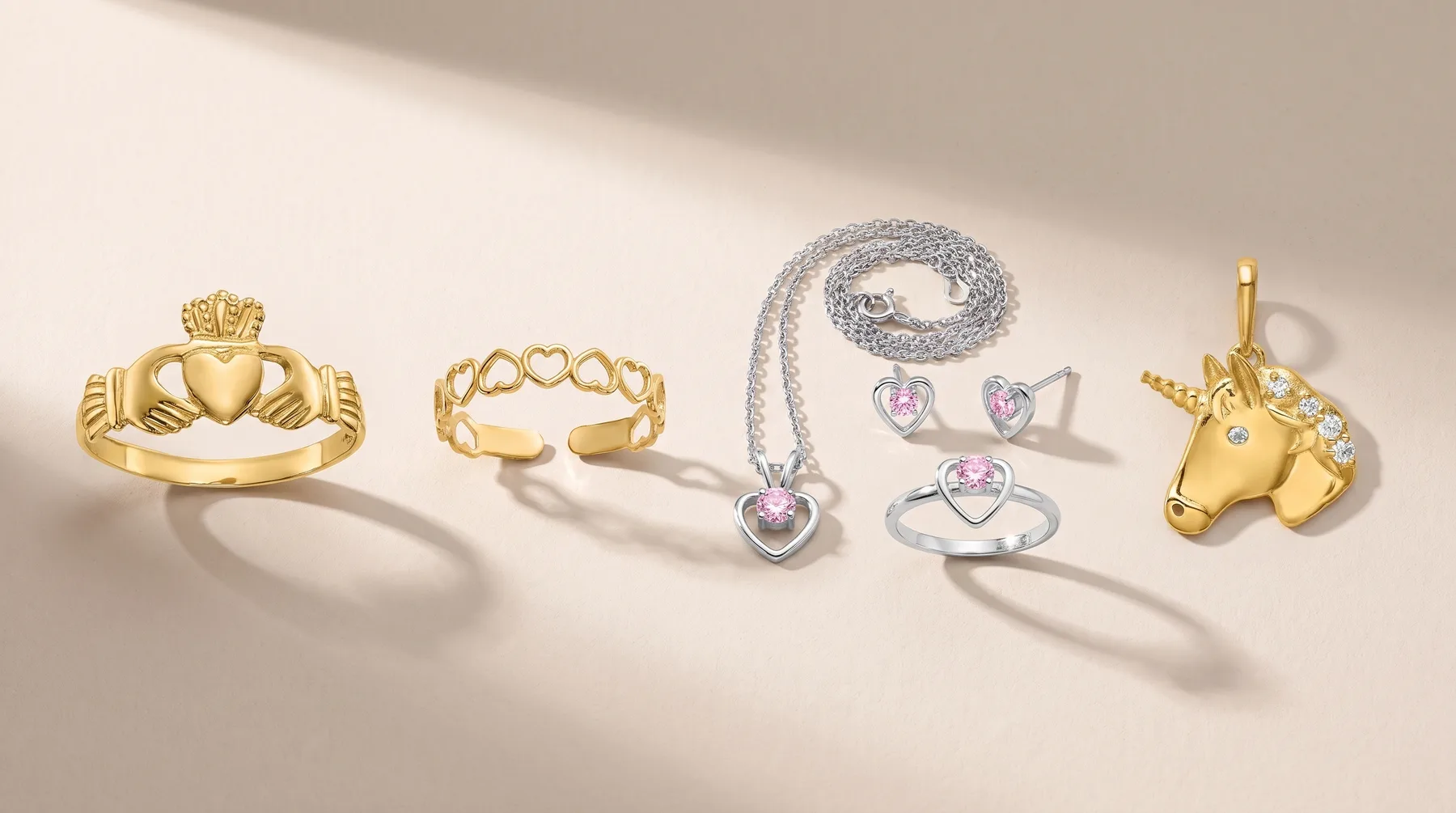 Five jewelry pieces displayed on a light background: a gold claddagh ring, a gold cuff bracelet, a silver necklace with purple heart-shaped stones, a silver ring with purple stones, and a gold unicorn head pendant.
