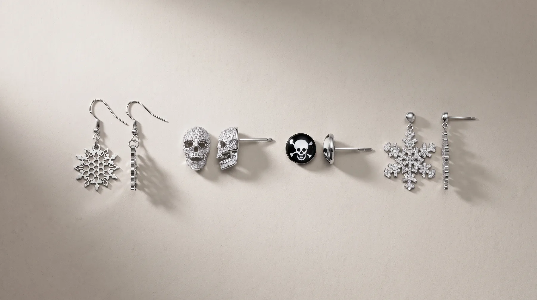 Six pairs of dangling earrings displayed in a row on a white surface, featuring snowflake designs, skull motifs, and stud styles in silver-tone metal with rhinestone accents.
