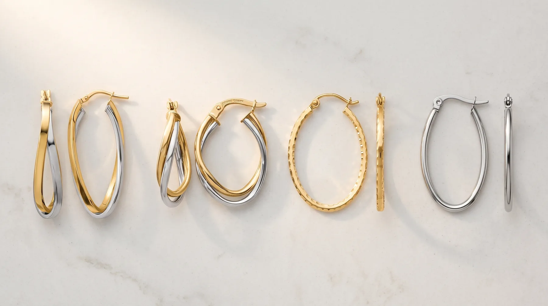 Seven pairs of hoop earrings in varying sizes and styles, including twisted, textured, and smooth finishes in gold and silver tones, arranged in a row on a marble surface.