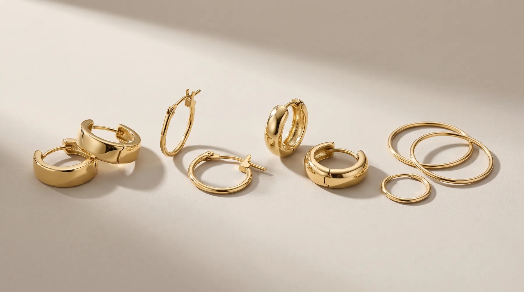 A collection of gold-toned hoop and band earrings in various widths and styles arranged on a neutral beige surface with soft shadows.
