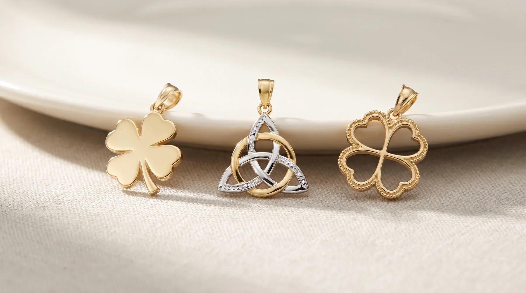 Three gold pendants displayed on a white plate: a four-leaf clover, a trinity knot with diamonds, and a heart-shaped four-leaf clover.