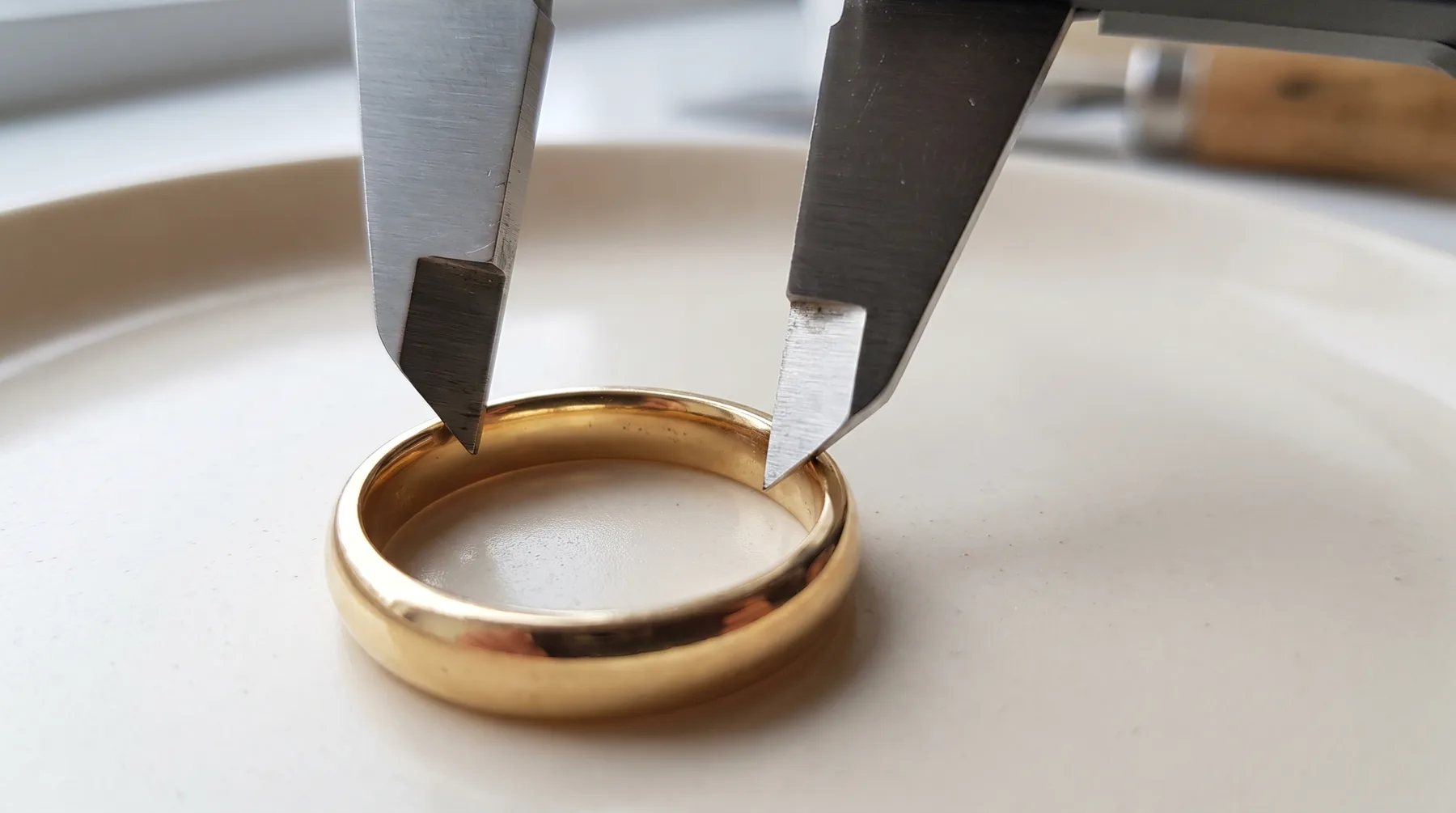 The inside jaws of a steel caliper spanning the interior of a plain gold band to measure its inside diameter, the ring resting on a pale ceramic dish.