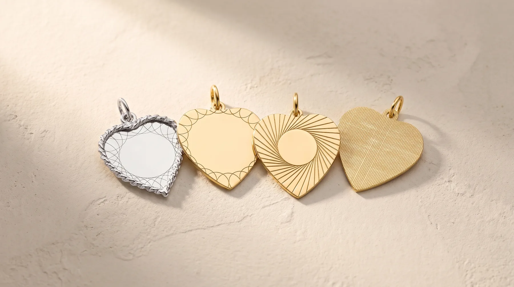 Four heart-shaped pendant charms in varying finishes arranged in a row: silver with textured border, polished gold, gold with radiating line pattern, and brushed gold.