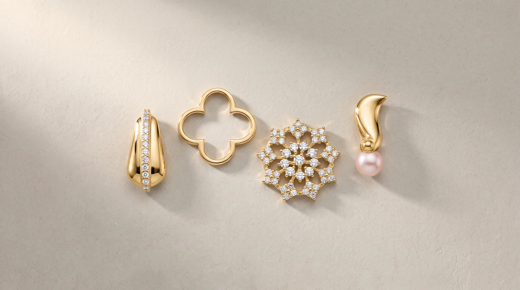 Four gold jewelry pieces arranged on a light surface: a textured oval pendant with diamonds, a four-leaf clover charm, a pearl and diamond cluster brooch, and a curved pendant with a pearl drop.