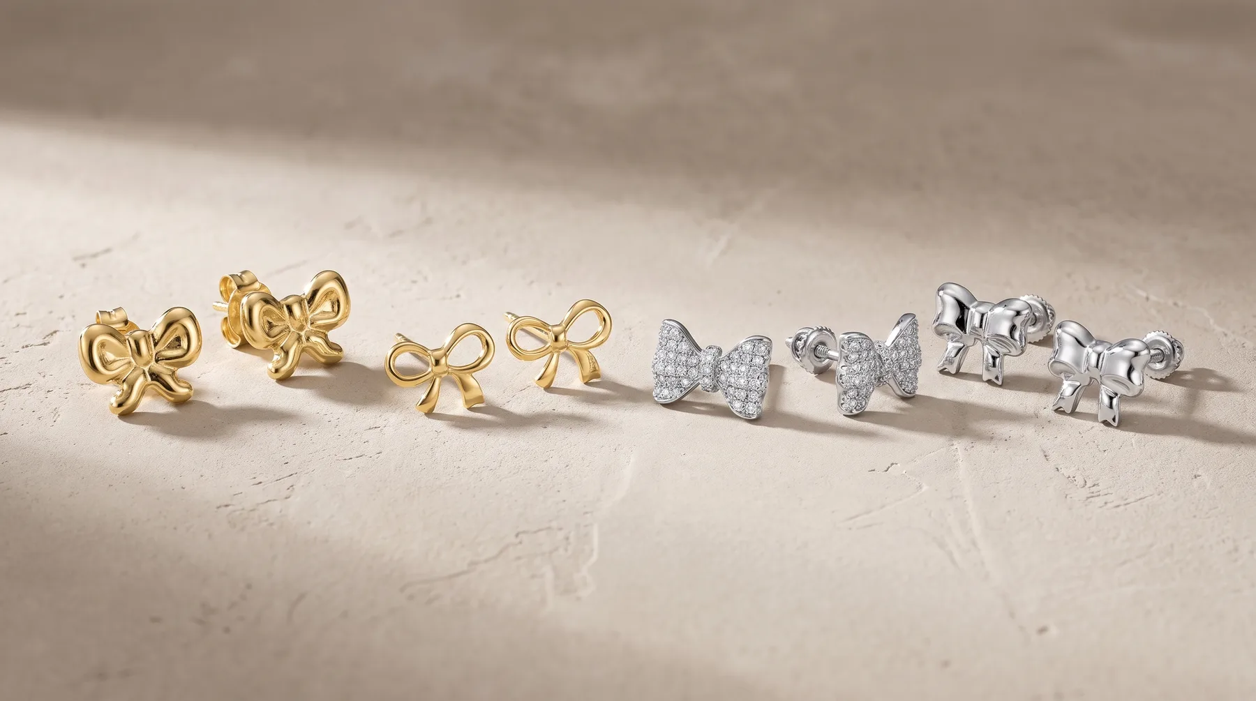 Six pairs of stud earrings arranged in a row on a textured beige surface: two gold bow-shaped pairs, two gray glittery bow pairs, and two silver elephant-shaped pairs.