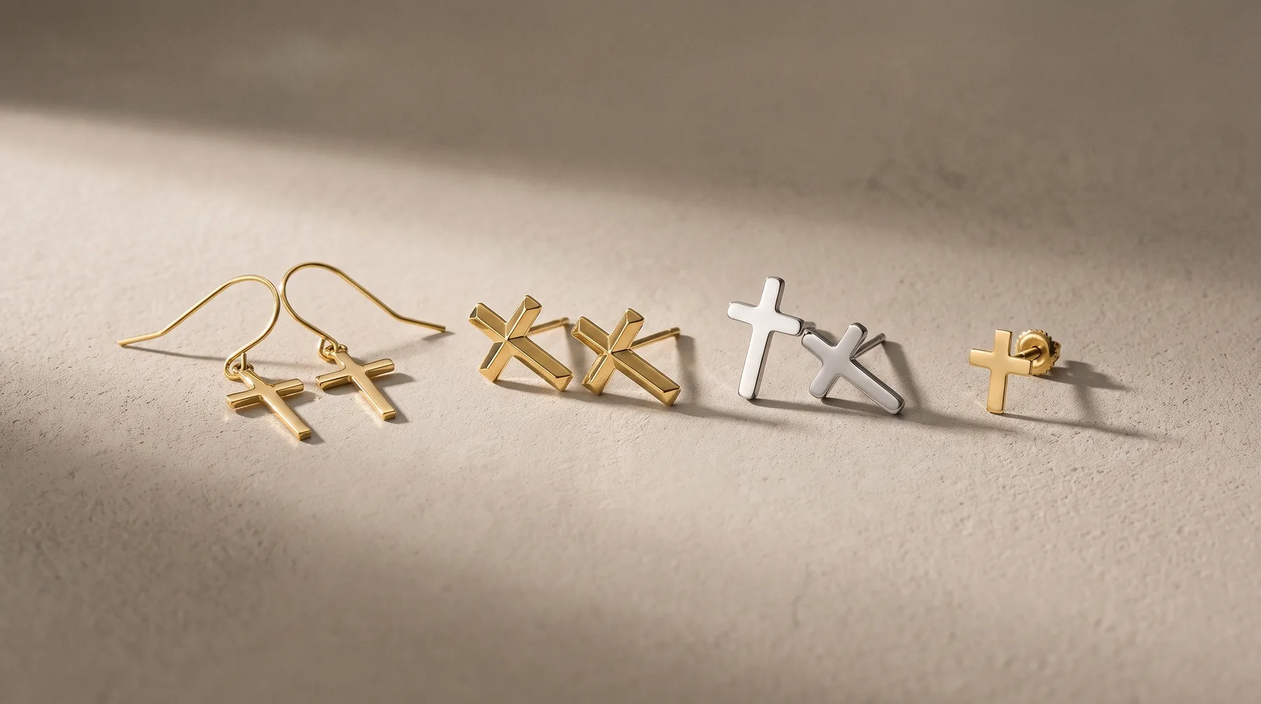Four cross-shaped jewelry pieces in gold and silver finishes arranged on a neutral surface, including a pair of drop earrings and two standalone pendants.