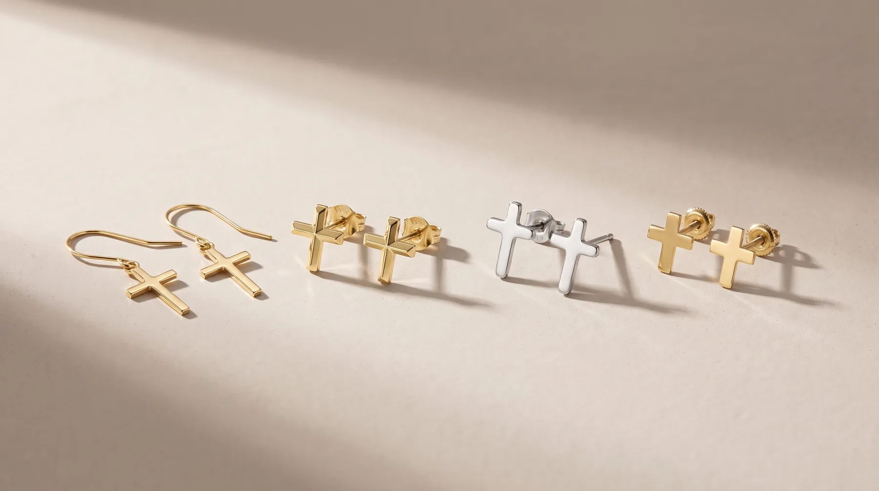 Five pairs of earrings arranged in a row on a white surface: two gold cross drop earrings, two gold gift box studs, two silver gift box studs, and two gold gift box studs.