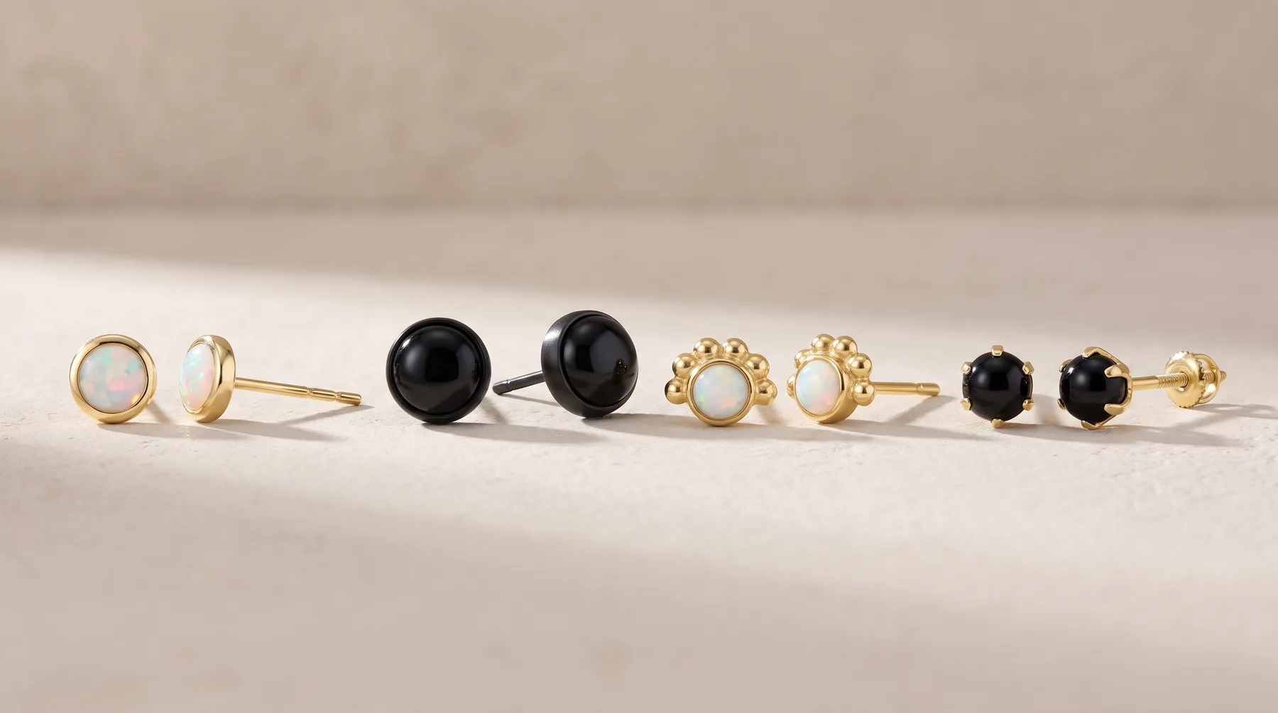 Eight gold stud earrings arranged in a row, featuring pearl, black onyx, and plain polished designs in various sizes.