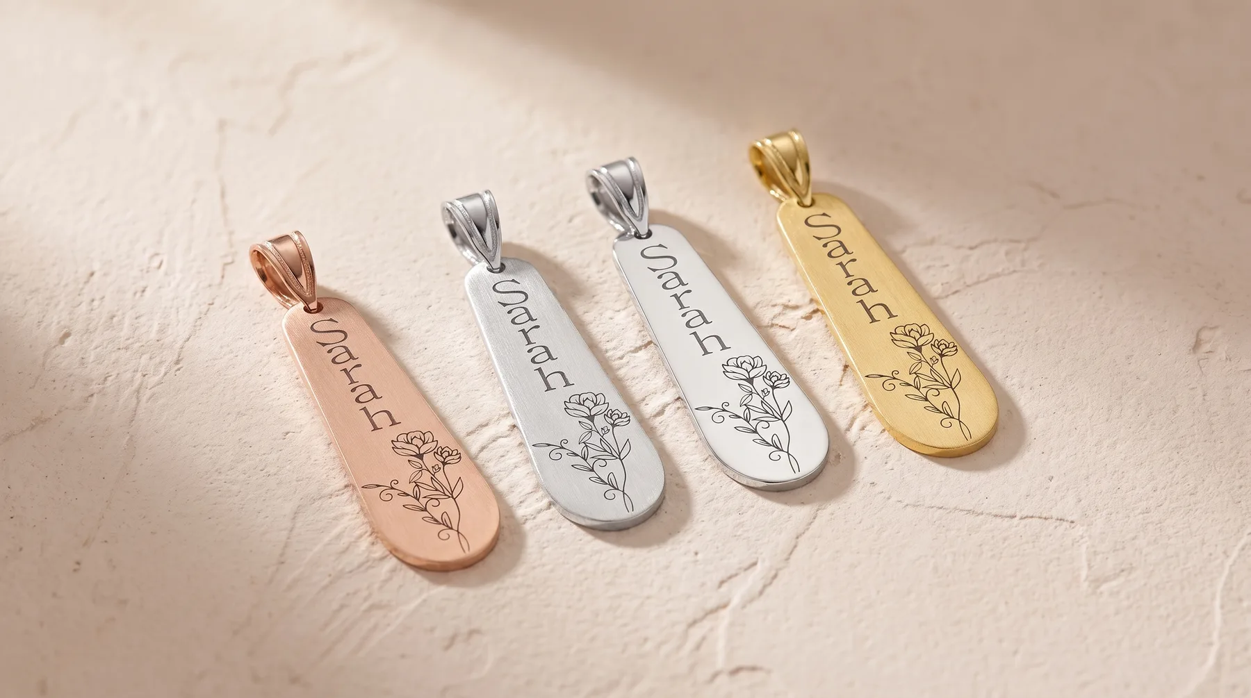 Four elongated teardrop-shaped pendants in rose gold, silver, and gold finishes, each engraved with text and a delicate botanical design, arranged in a row on a textured beige surface.