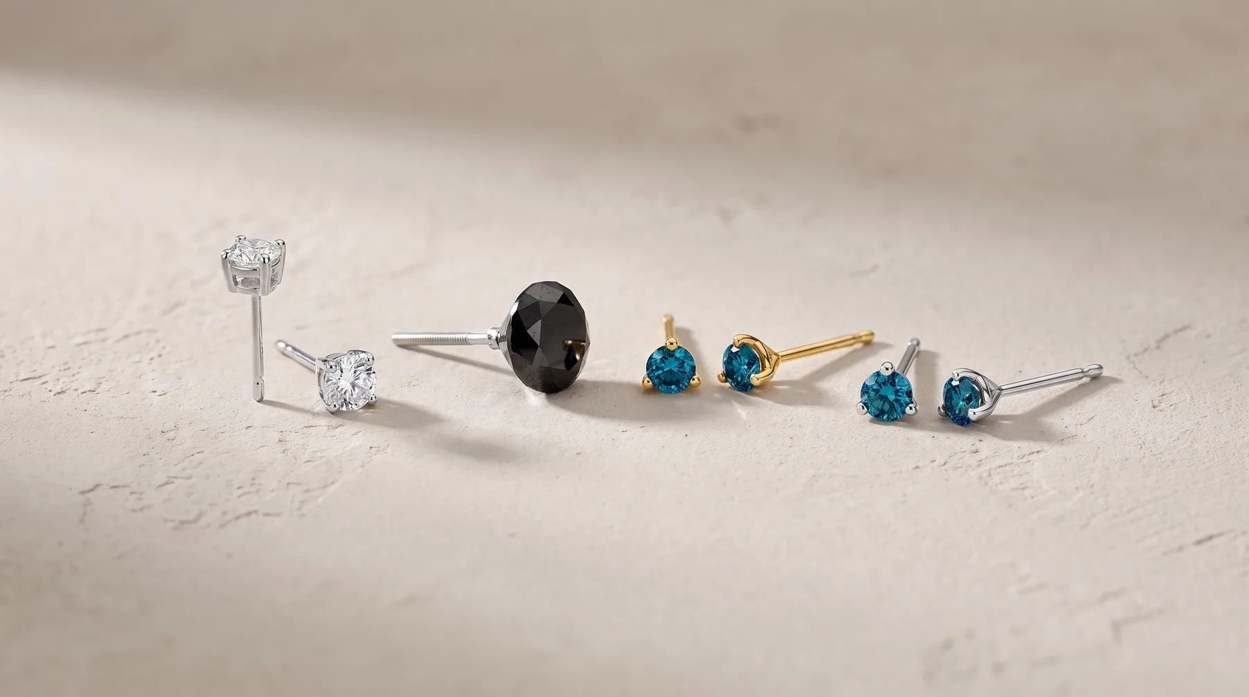 Six stud earrings arranged in a row on a light marble surface, featuring clear and blue gemstones in silver and gold settings.