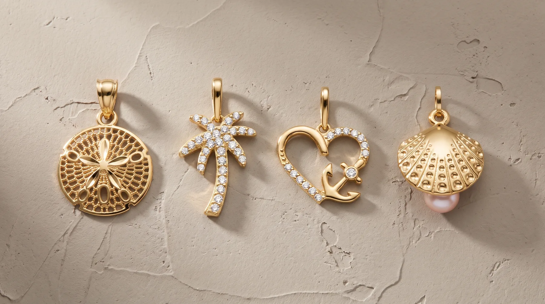 Four gold pendant charms displayed on a textured beige surface: a circular filigree disc, a palm tree with crystal accents, a heart with a pearl drop, and a scallop shell with a pearl.