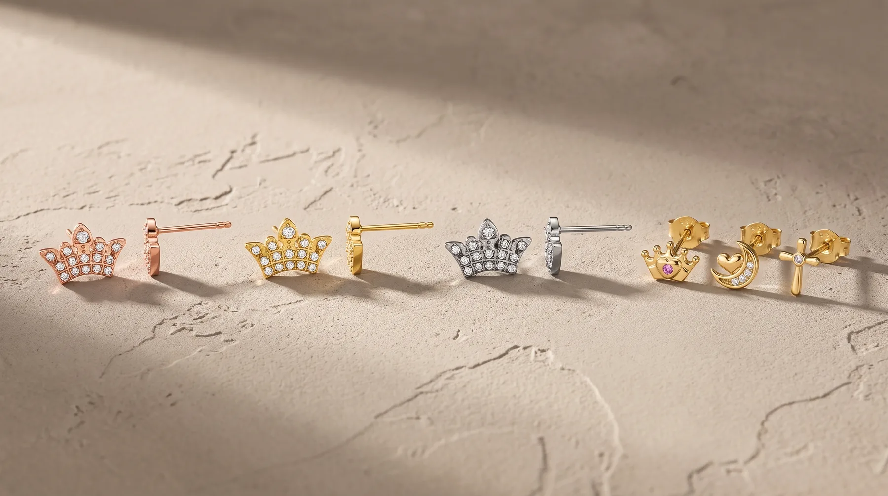 A collection of crown-shaped stud earrings in various metals including rose gold, yellow gold, and silver, displayed in a line on a textured beige surface.