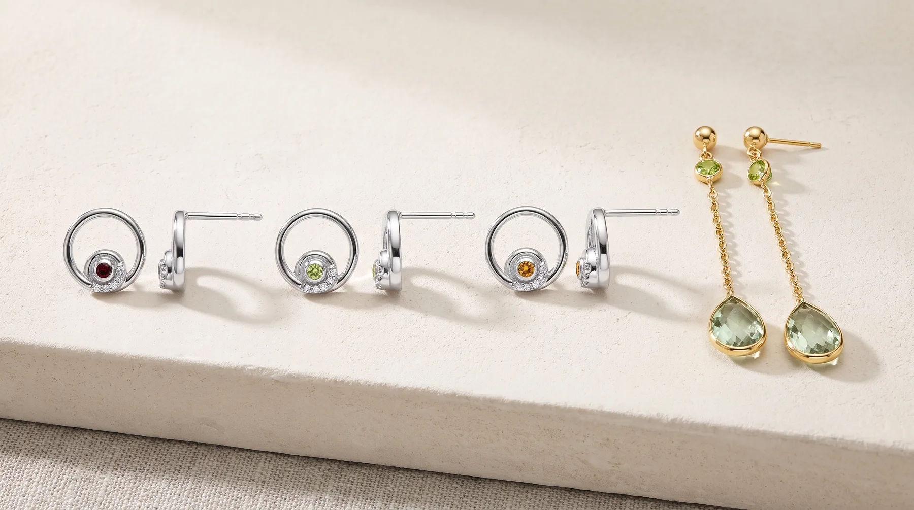 Five pairs of gemstone earrings displayed on a white rectangular platform, including three silver circular designs with colored stones and two gold chain drop earrings with faceted pale green stones.