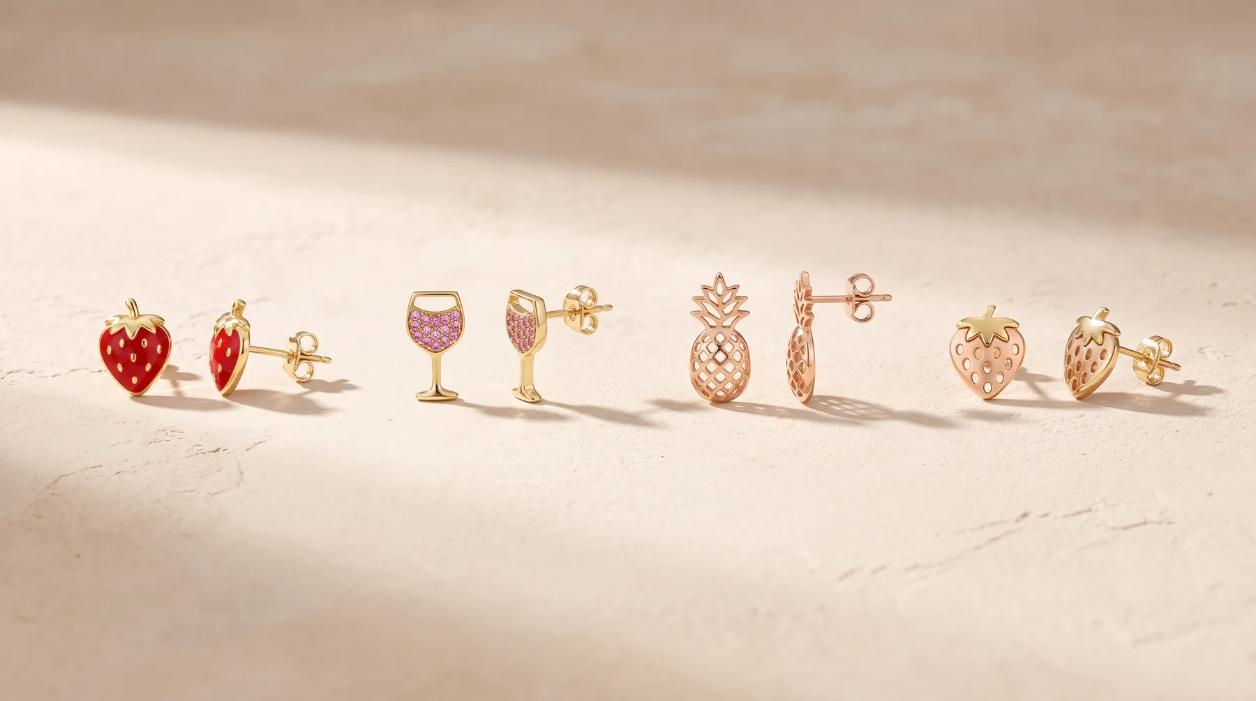 Five pairs of gold stud earrings with fruit designs—strawberries, wine glasses, pineapples, and hearts—arranged in a row on a neutral surface.