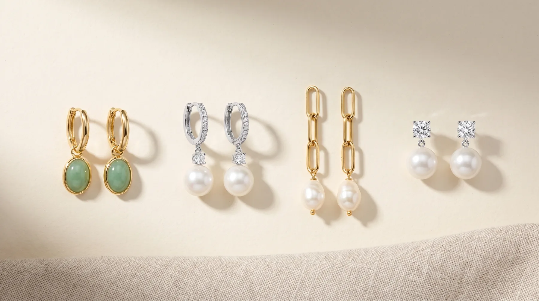 Four pairs of earrings arranged in a row on a beige surface: gold hoops with green stone drops, silver hoops with white pearl drops, gold chain-link style with white pearl drops, and gold studs with white pearls and diamond accents.