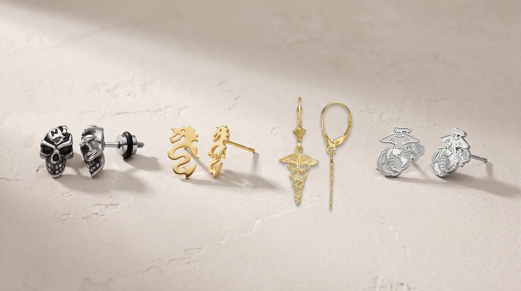 Four pairs of earrings and two pendant necklaces arranged in a row on a light textured surface: skull studs in silver, wavy gold studs, a gold sword pendant on a chain, a gold loop pendant on a chain, and eagle studs in silver.