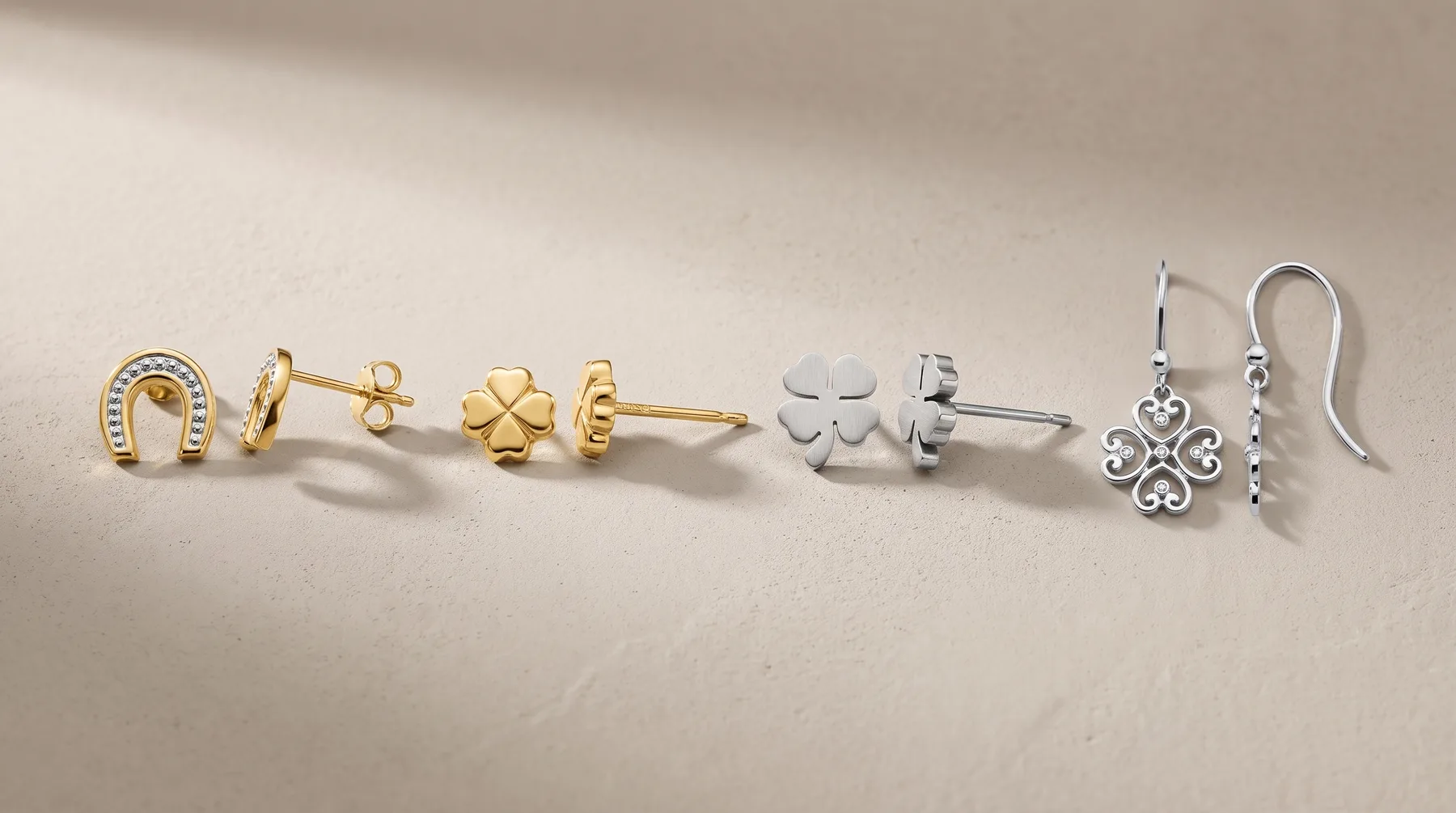 Five pairs of earrings displayed in a row on a beige surface: gold horseshoe studs, gold four-leaf clover studs, silver four-leaf clover studs, and silver ornate drop earrings with a fish hook backing.