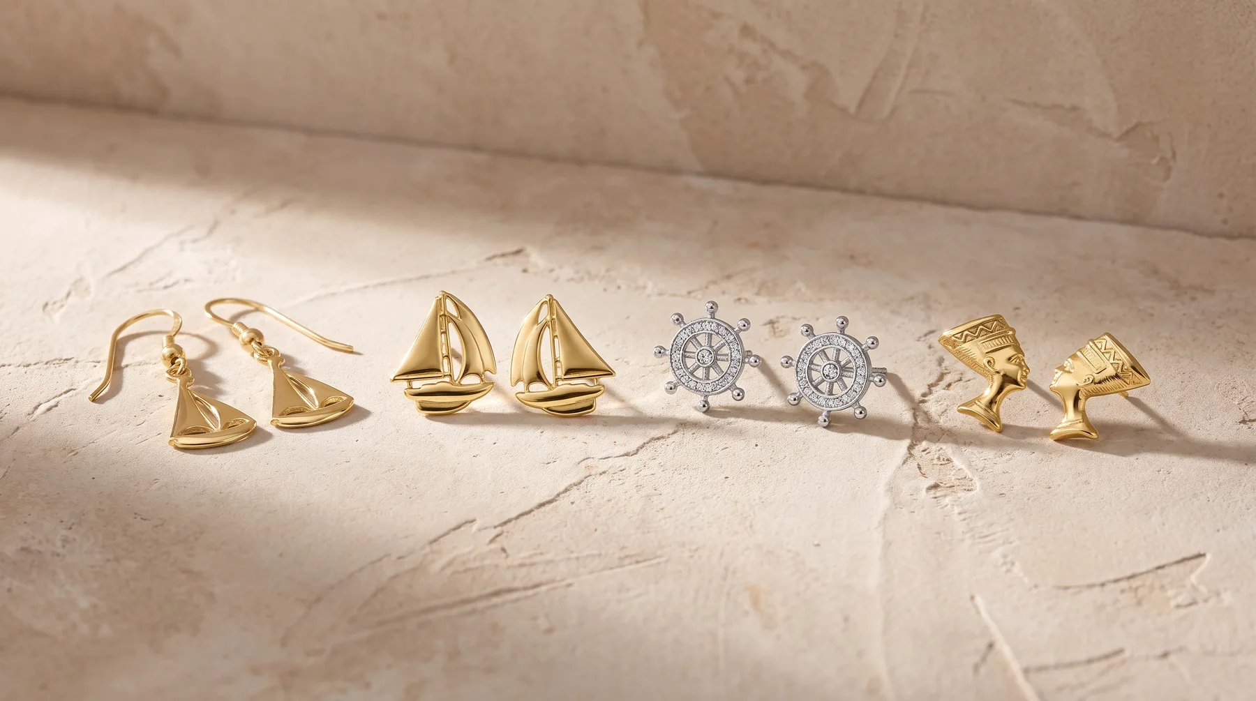 Six pairs of earrings arranged in a row on a marble surface, including gold sailboat designs, silver ship wheel charms, and gold pyramid studs.