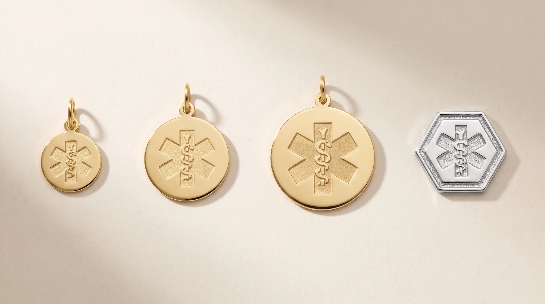 Four medical alert pendants arranged in a row: three gold-colored circular discs with the Rod of Asclepius symbol in decreasing sizes, and one silver-colored hexagonal pendant with the same symbol.