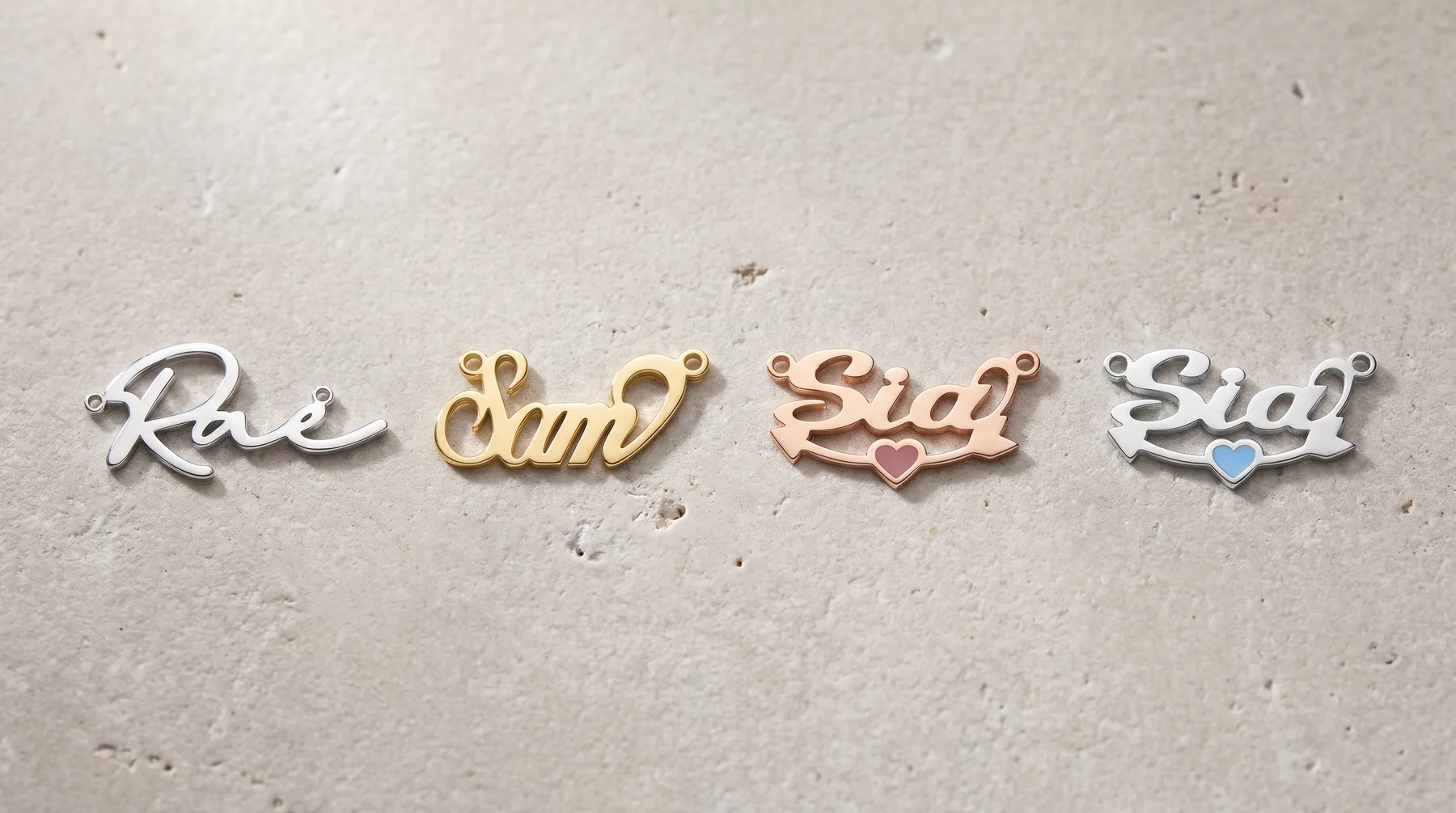 Four metal charms in different finishes arranged in a row on a concrete surface, each featuring cursive text with decorative elements and small gemstones.
