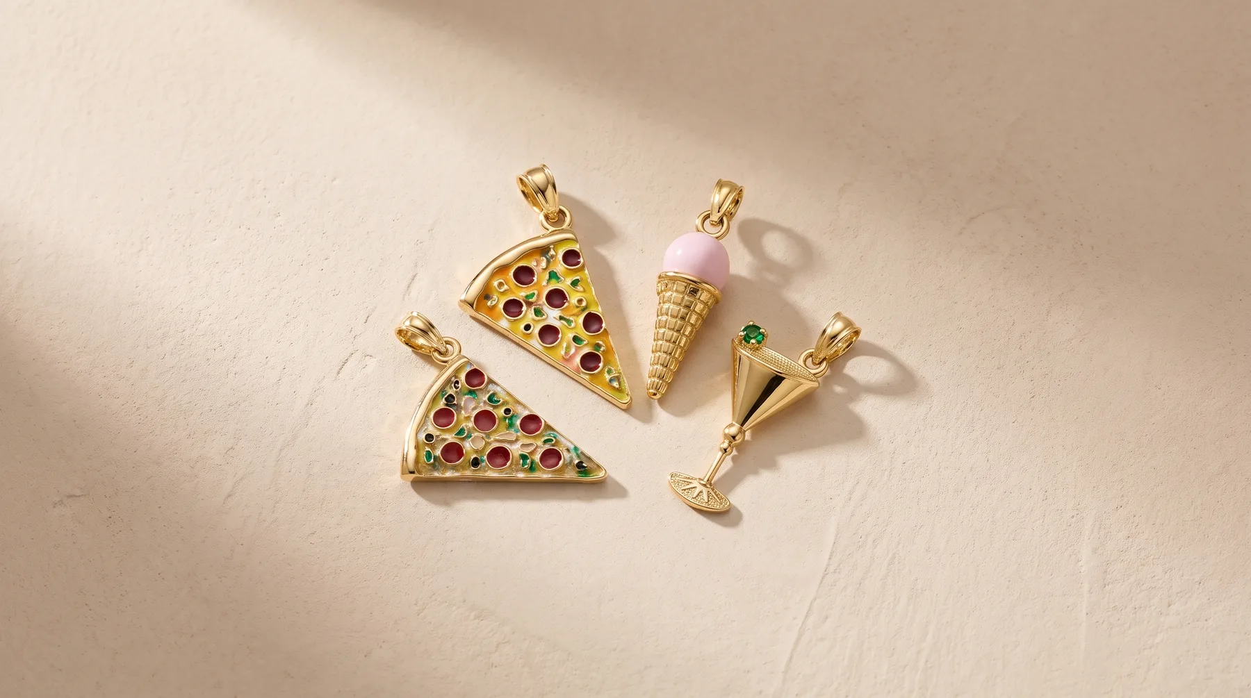 Four gold-toned charm pendants arranged on a beige surface: two triangular pizza slices set with colorful gemstones, one cone-shaped ice cream charm, and one martini glass charm with a small hallmark.