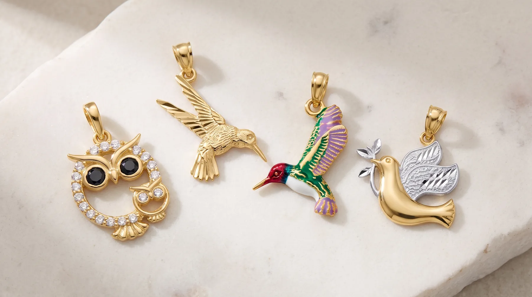 Four gold charm pendants displayed on a marble surface: an owl with black onyx eyes and crystals, a hummingbird, a colorful parrot with green and pink enamel, and a dove with an engraved circular disc.