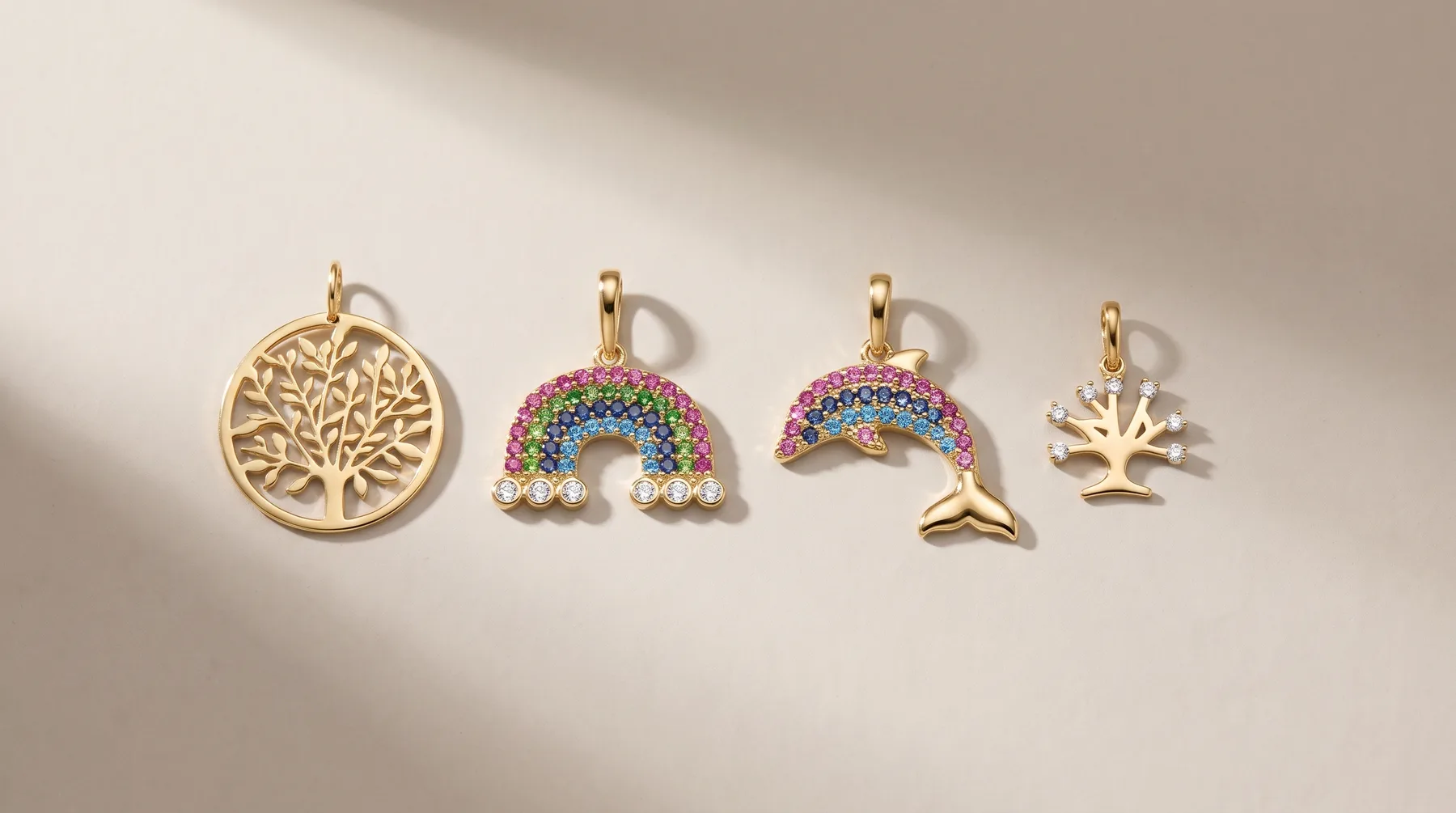 Four gold pendant charms arranged in a row: a circular tree of life design, two rainbow-shaped pendants with colorful gemstones, and a small crown or leaf-shaped charm.