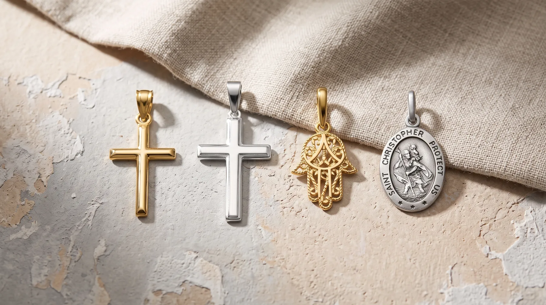 Four religious pendants with bails arranged in a row on a textured beige surface with draped cream fabric: two crosses in gold and silver, a gold hamsa hand, and a silver oval medallion with a saint figure.