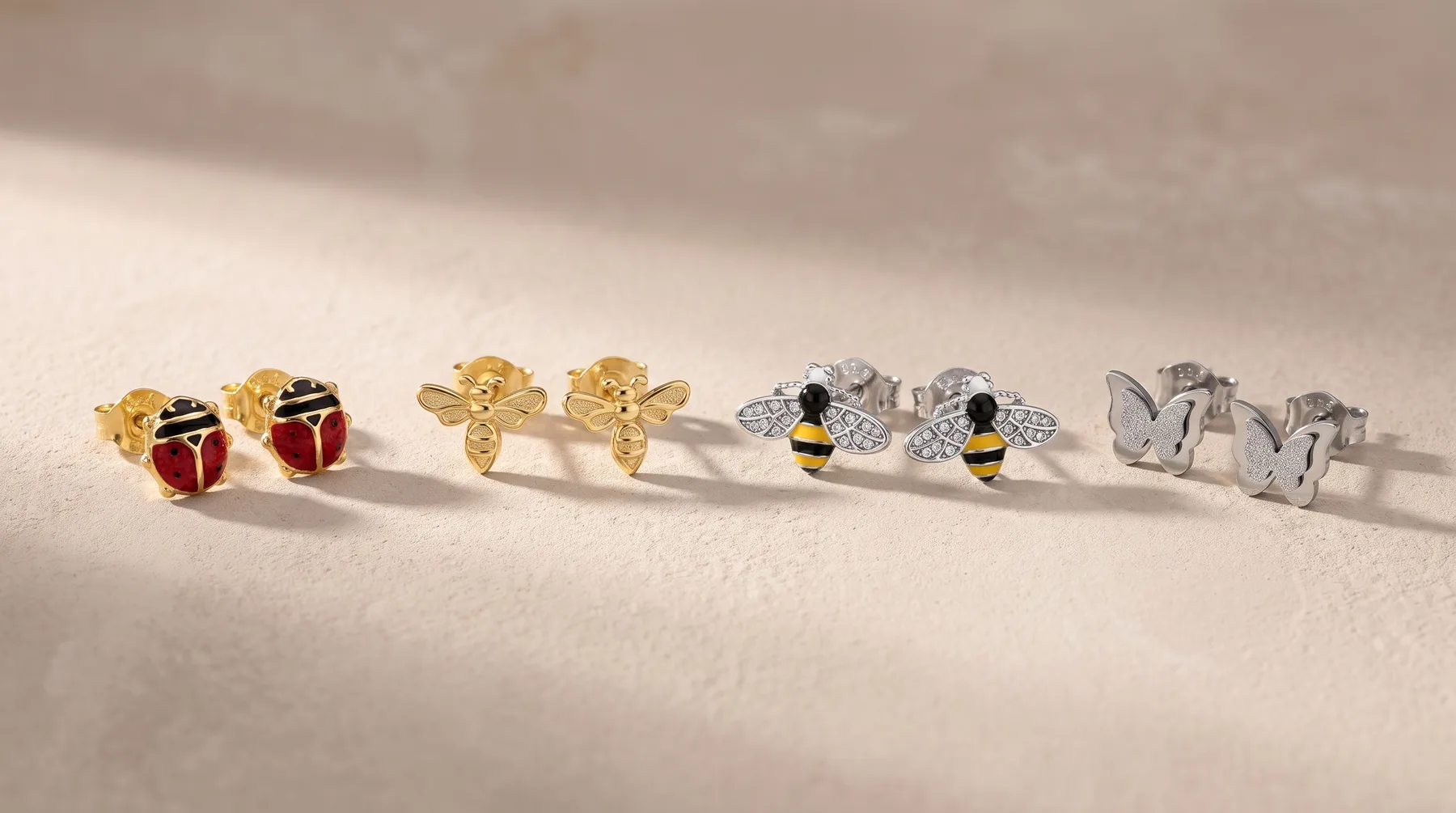 Four pairs of stud earrings arranged in a row on a light surface: red and black ladybug designs in gold, gold flowers, silver bees with black and yellow stripes, and silver elephant heads.