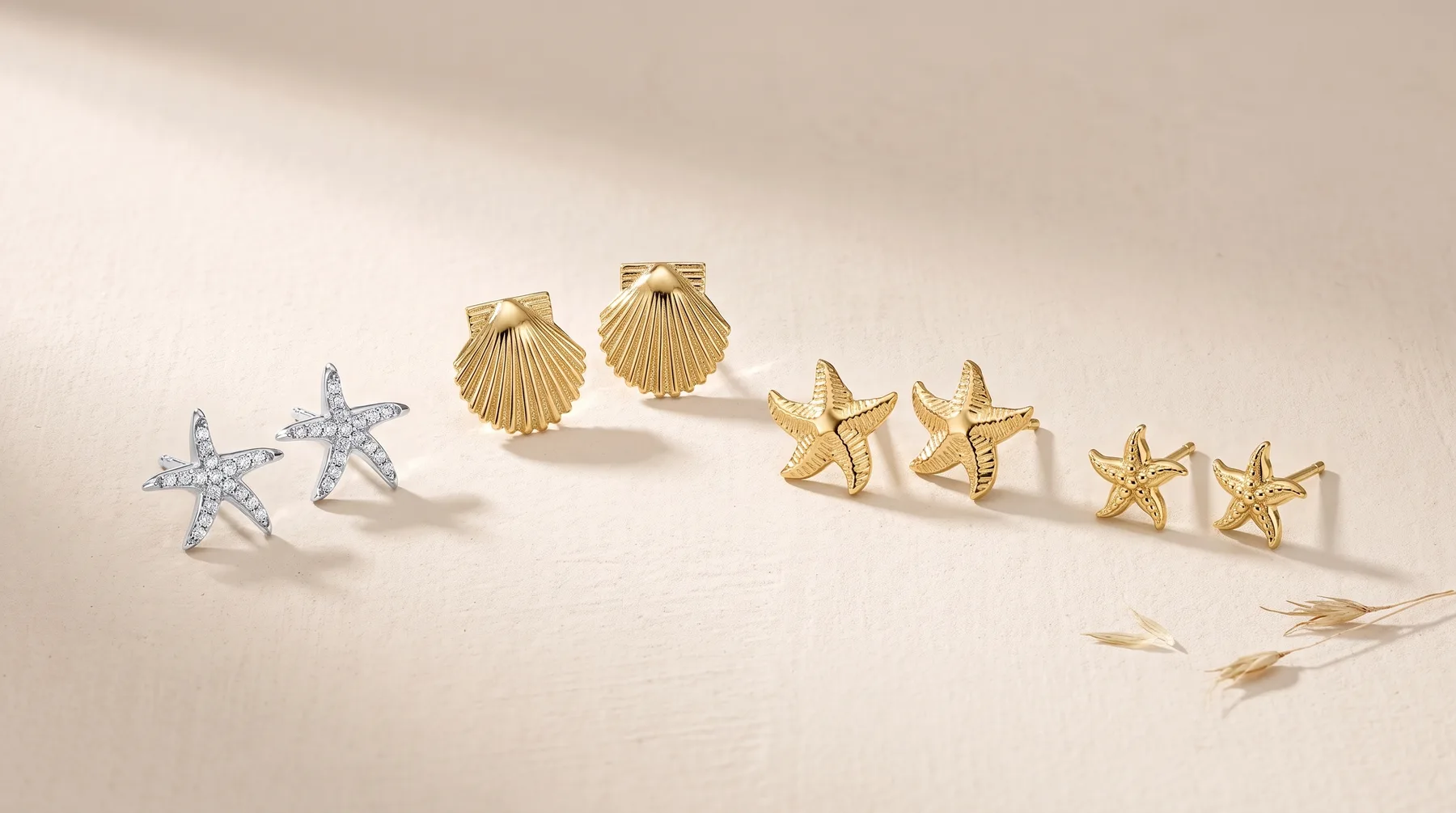 Six pairs of earrings arranged in a line on a neutral surface: two silver textured starfish studs, two gold shell studs, and two pairs of smaller gold starfish studs, with dried grasses nearby.