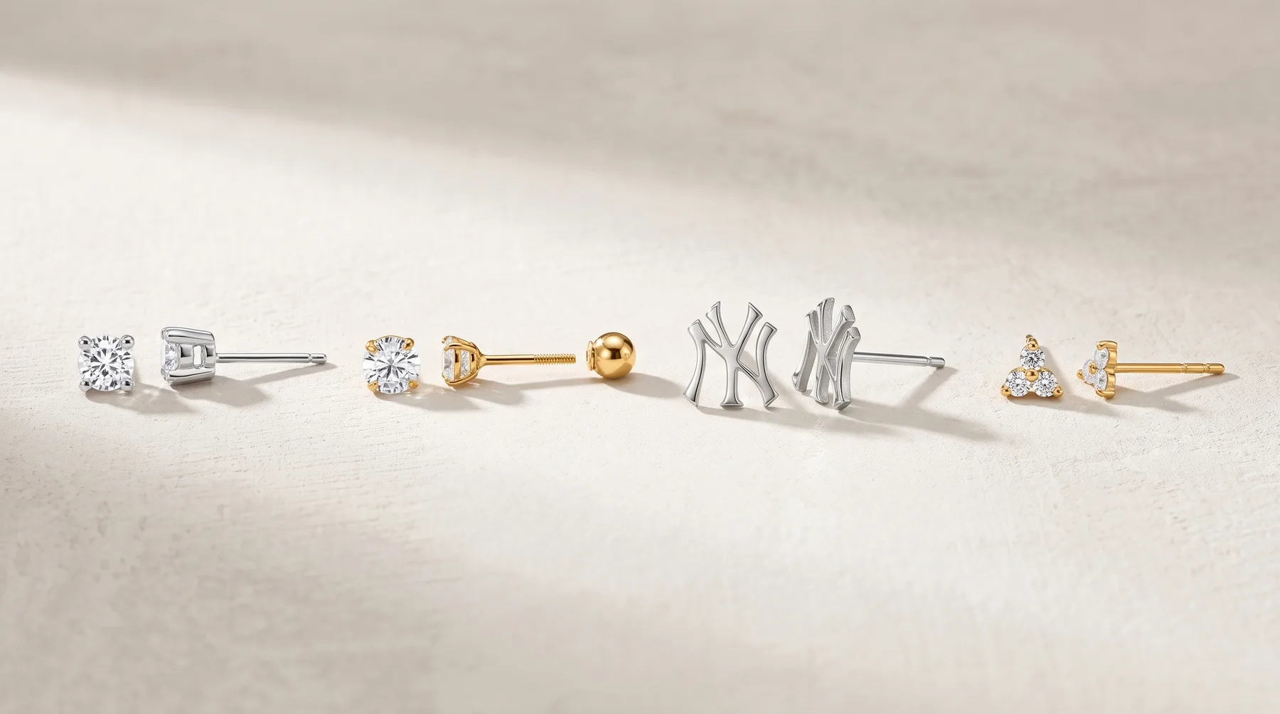 Five pairs of stud earrings arranged in a line on a light stone surface, including diamond studs in white and yellow gold, a Yankees logo pair, and small gemstone studs.