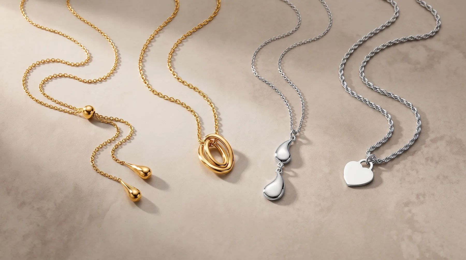 Three delicate chain necklaces in gold and silver finishes displayed on a neutral surface, each featuring small pendant charms including spheres and heart shapes.