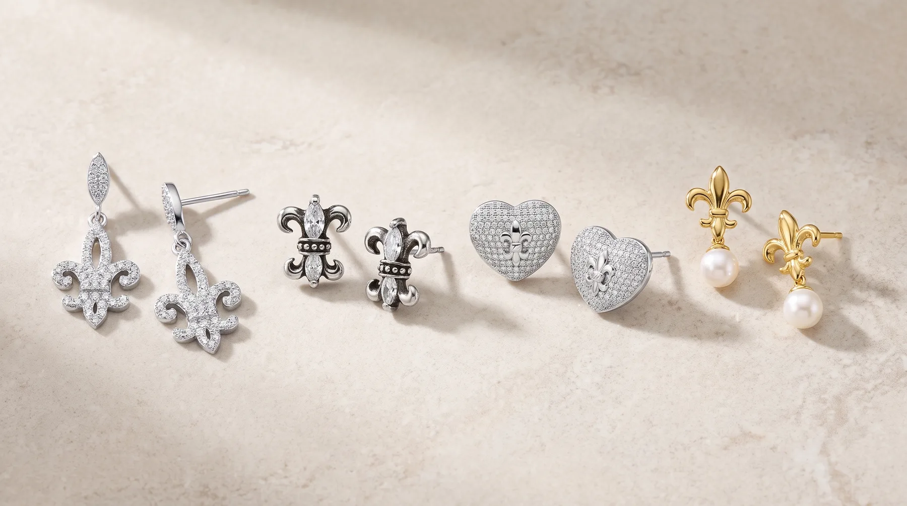 Four pairs of earrings arranged in a row on a marble surface: diamond fleur-de-lis drops in silver, ornate fleur-de-lis studs in silver and black enamel, heart-shaped diamond studs in silver, and gold fleur-de-lis studs with pearl drops.