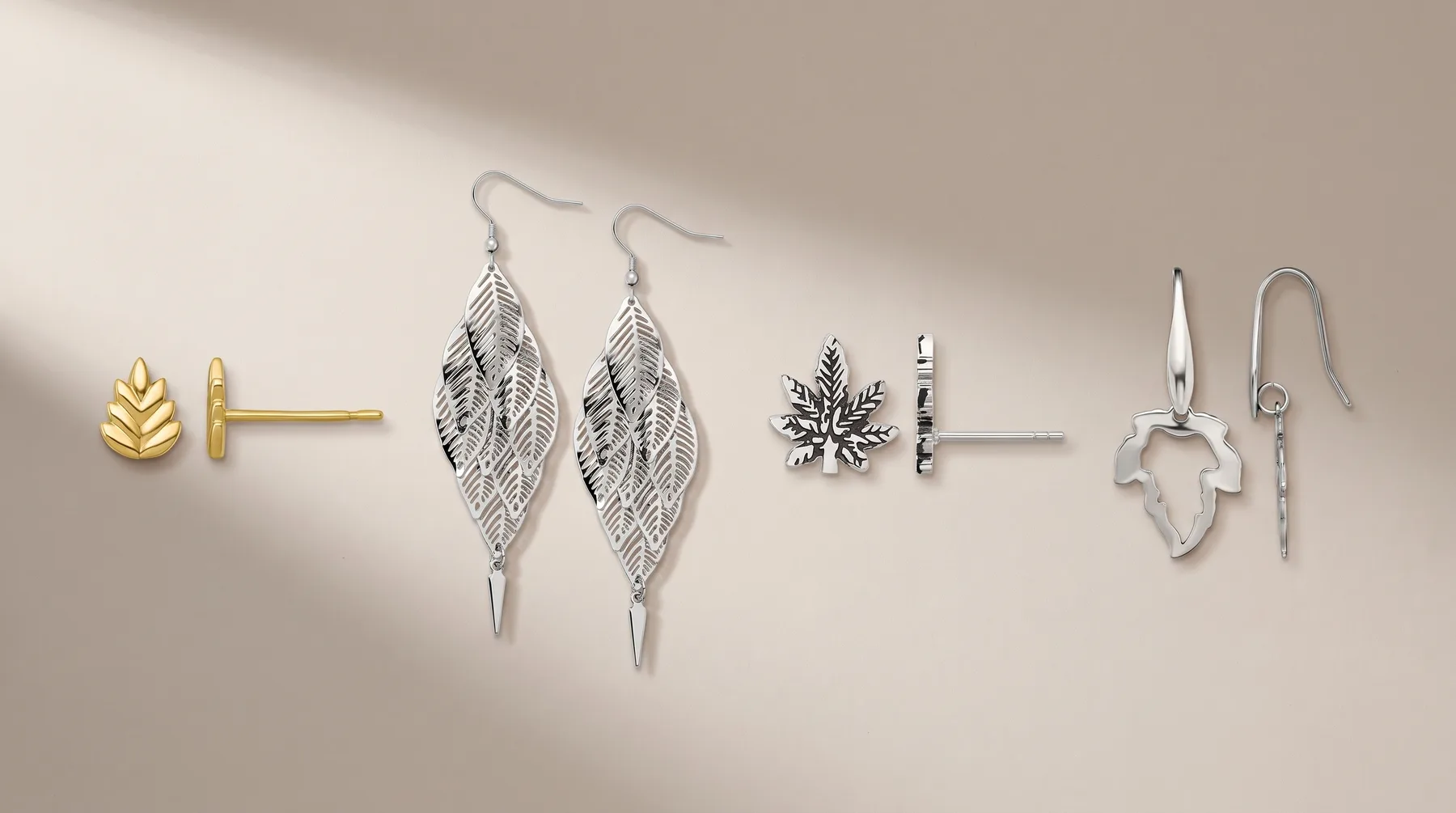Six pairs of earrings displayed on a beige surface, including gold leaf studs, silver filigree leaf drops, a snowflake design, black striped bar studs, and silver abstract shape drops.