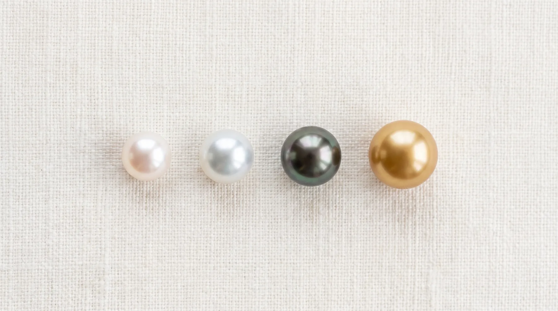 Four loose cultured pearls in a row on ivory linen, increasing in size from a small rose-white pearl to a silvery white one, a dark peacock-grey pearl, and a large deep golden pearl